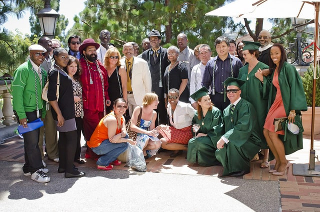 A large, diverse group of people gathered outdoors, smiling and posing together on a sunny day. Four individuals in the front wear green graduation caps and gowns, while others are dressed in a mix of casual and semi-formal attire. The group stands on a brick patio surrounded by greenery, trees, and outdoor umbrellas, suggesting a celebratory occasion like a graduation or ceremony.